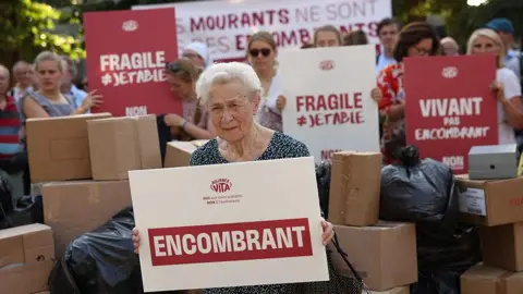 AFP via Getty Images A protester sits with a placard that reads "Burden" during a rally organised by French "pro-life" organisation Alliance Vita against the new French draft law on euthanasia and assisted suicide at Invalides, in Paris on June 22, 2026
