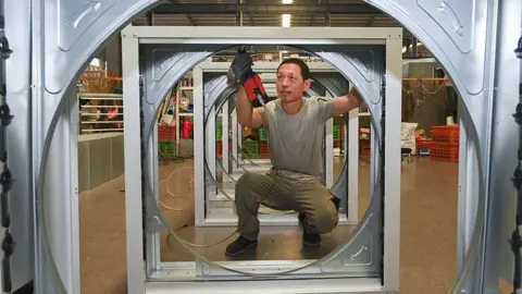 VCG via Getty Images An employee at a wind turbiner manufacturing company uses work tools to put together some steel parts. In the image, he is pictured squatting at the centre of one of the steel frames.