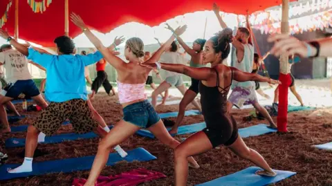 Josh Harkon Women and men wearing shorts and T-shirts are standing on yoga mats on grass underneath a red canopy