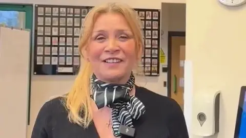 LDRS Cathy Hunt, a middle-aged woman with mid-length blonde hair, wearing a black neckerchief and a gold necklace with a cross pendant. She has a black blouse and stands in front of a grid showing small head and shoulders images of people.