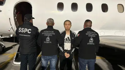 Secretary Omar Harfuch of Mexico's Security and Citizen Protection Zhang Zhidong standing in front of a plane in handcuffs. He is wearing a dark blue tracksuit and white Adidas top. He is facing the camera, while three police officials standing beside him have their backs turned to the camera.