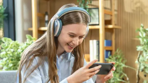 Getty Images A woman with long light brown hair playing a game on her phone in a living room while wearing blue headphones and a blue shirt