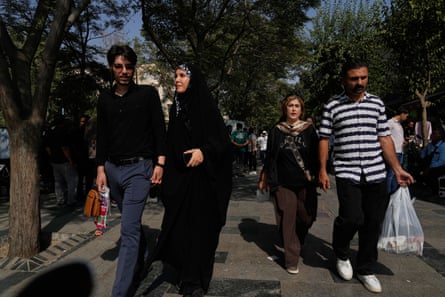 Two couples walk down a tree-lined outdoor path near a market. One man holds a plastic shopping bag. They appear to be quite affluent-looking and well-dressed city dwellers. One of the women wears a headscarf.