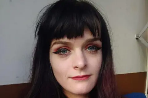 Abi Smith A selfie of a woman who has long, dark hair including a fringe. She is sitting on a sofa and is wearing a dark navy blue jumper.