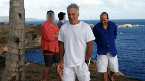 US Department of Justice Several people stand outdoors beside a coastline overlooking the sea. In the foreground, Jeffrey Epstein in a white T-shirt and Howard Lutnick in a blue button-down shirt are visible, with rocky outcrops and deep blue water in the background under a cloudy sky. Two other people, blurred, stand to the left.
