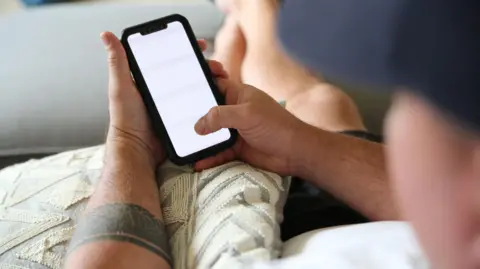 Getty A man with a tattoo on his arm looks at a white mobile phone screen
