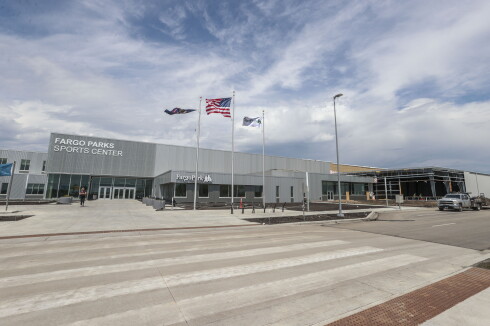 Seen from a crosswalk, a low-rise silver building sits beneath cloudy skies. Signage indicates it is the "FARGO PARKS SPORTS CENTER." A portion of the building is still under construction at right. Wind whips three flags on poles in front of the building.