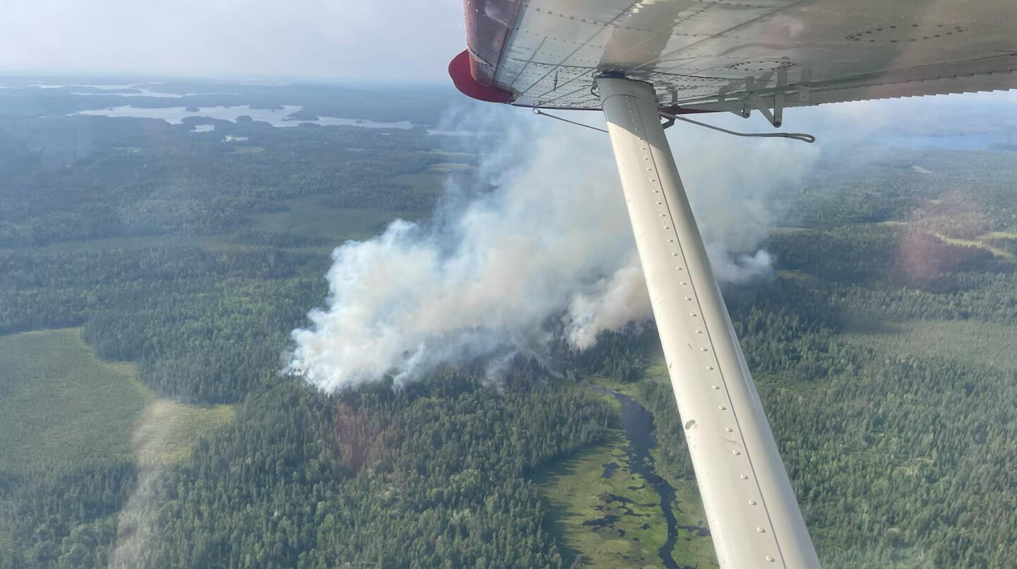 Smoke rises from a forest in an aerial photograph. A plane's wing is visible