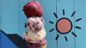 A hand holds three colorful scoops of ice cream — a mix of deep purple and berry shades — in front of a blue mountainous mural.