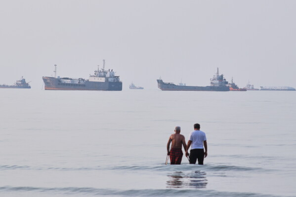 Two men wade in the waters of the Strait of Hormuz with vessels anchored in the background, off Bandar Abbas, Iran, Sunday, July 12, 2026. (Razieh Poudat/ISNA via AP)