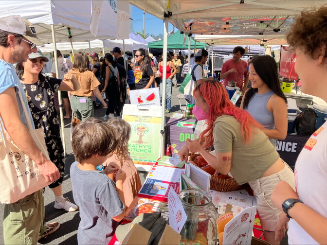 A woman with a mask and orange hair leaning showing children a book under at a tent in a farmers' market.