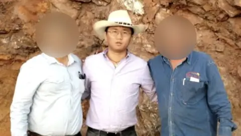 'Alex' A man wearing a white cowboy-style hat and a pale purple shirt tucked into his trousers. He is wearing glasses and a necklace and is looking at the camera. There is a man on his left and another on his right - the faces of these two men  are blurred to obscure their identities.