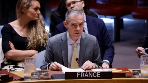 Getty Images France's UN Ambassador Jerome Bonnafont speaking at the UN Security Council on the table with a "France" placard while two people sit behind him
