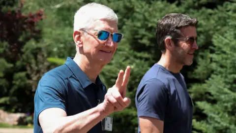Reuters Apple CEO Tim Cook walking outside, throwing up 'peace' fingers, wearing a blue polo shirt and reflective sunglasses. John Ternus, who will replace Cook as CEO later this year, walks beside him, wearing a blue t-shirt and sunglasses.