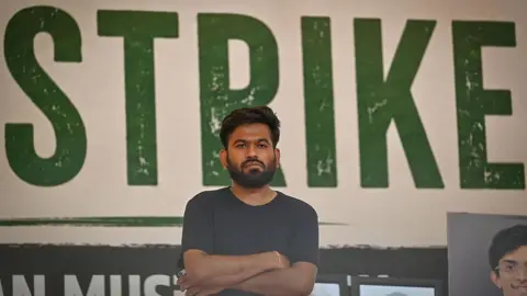 Getty Images CJP founder Abhijeet Dipke wearing a black T-shirt with his arms crossed. Behind him is a large poster where the word 'STRIKE' can be seen