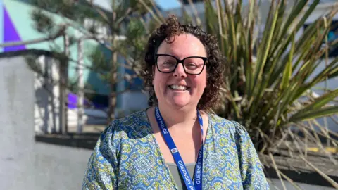 BBC A woman with curly brown hair and black glasses. She is wearing a blue and green top with a light green vest. She is wearing a blue lanyard which says "Guernsey Police" on. Behind her is a white wall and green bush.