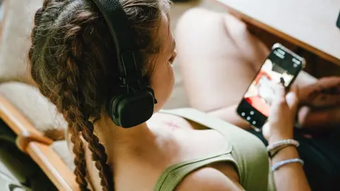 Getty Images A close-up, over-the-shoulder stock photo shows a young person relaxing in a wooden chair while using a smartphone. The person wears large black over-ear headphones and has long hair styled in two braids. A light green sleeveless top is visible, along with bracelets on one wrist. The smartphone is held in one hand and displays a media or video-related screen, although the content is slightly out of focus.