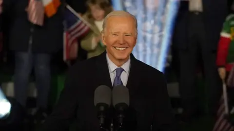 PA Media A man with white hair smiles. He is standing at a podium with an American eagle design on the front. He is wearing a suit. Behind him is a crowd, with a mixture of Irish and American flags.