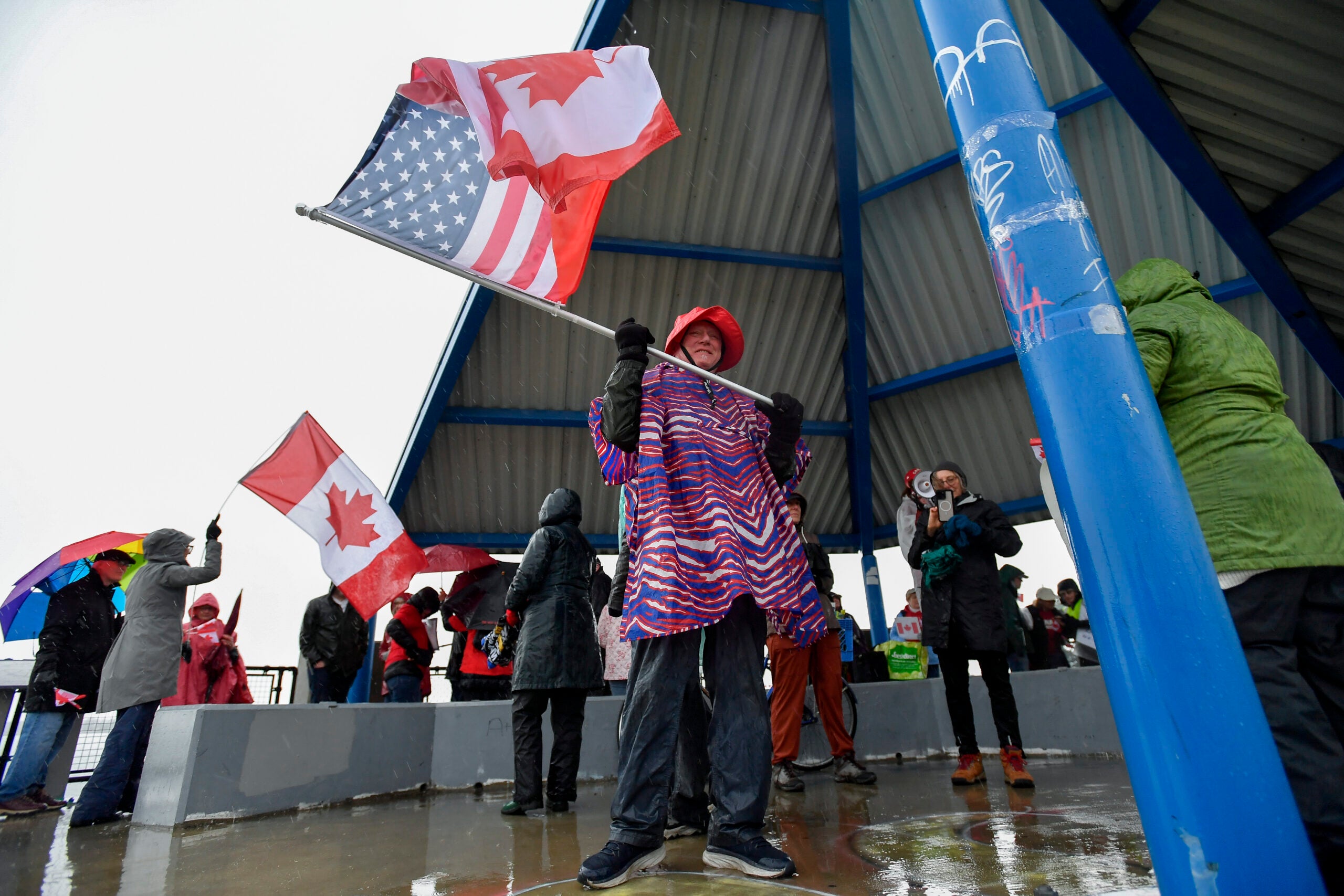 People gathered at an outdoor pavilion in raincoats holding Canadian and American flags on a rainy day.