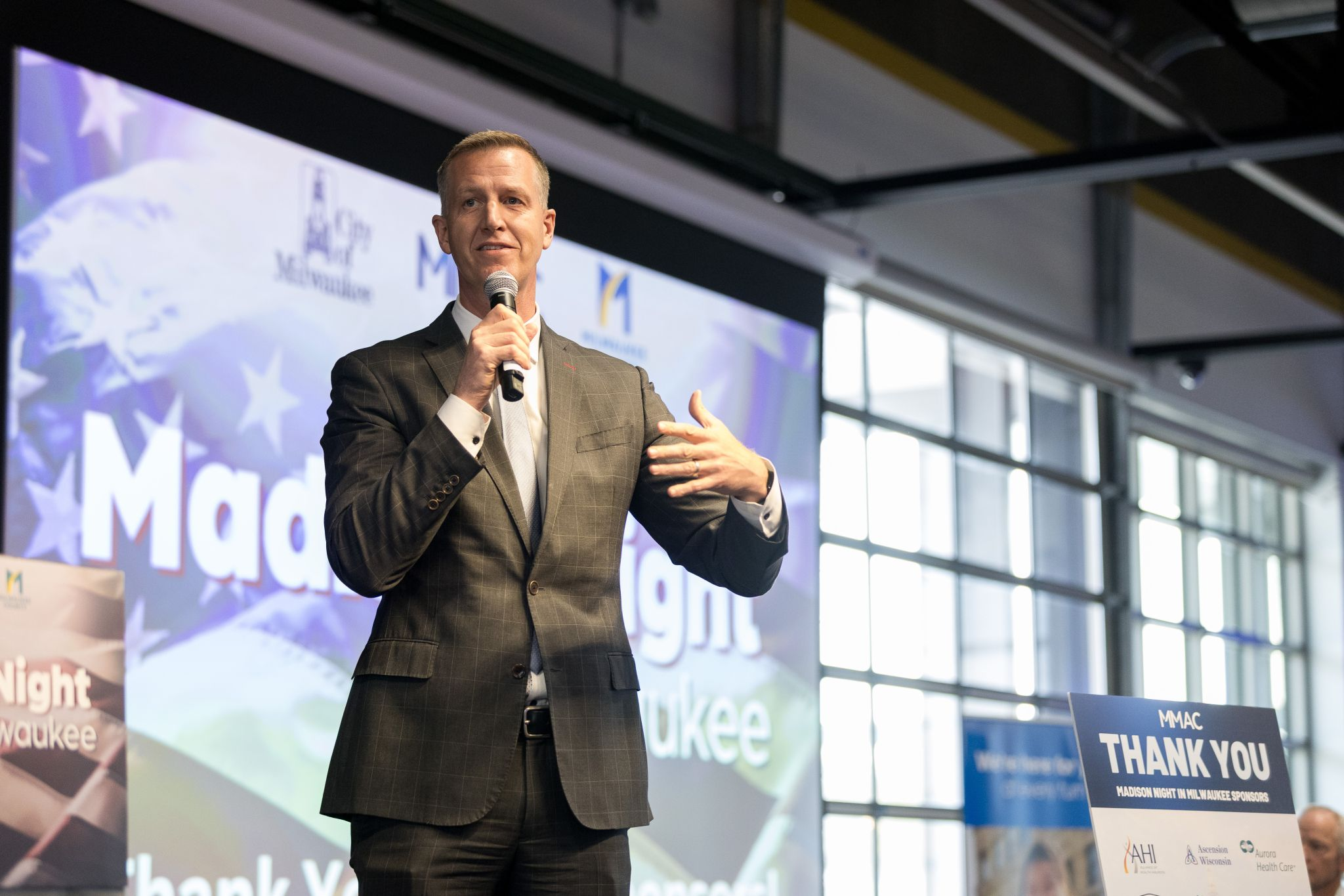 A man in a suit speaks into a microphone onstage in front of a screen displaying Madi...light waukee at an indoor event with large windows.