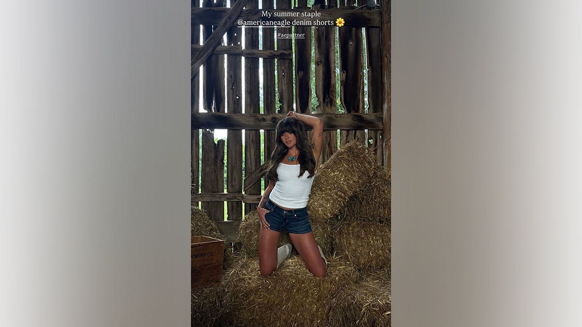 Ella Langley kneels atop stacked hay bales while wearing a white tank top, dark-wash denim shorts and white cowboy boots during an American Eagle photoshoot.