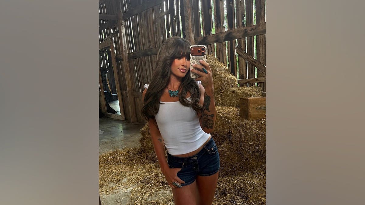 Ella Langley smiles while taking a mirror selfie in a white tank top, dark-wash denim shorts and white cowboy boots inside a rustic barn.