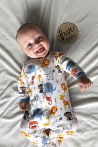 A baby boy smiles as he lies on a blanket wearing a onesie with an animal print. A sign next to him reads “Five months.”