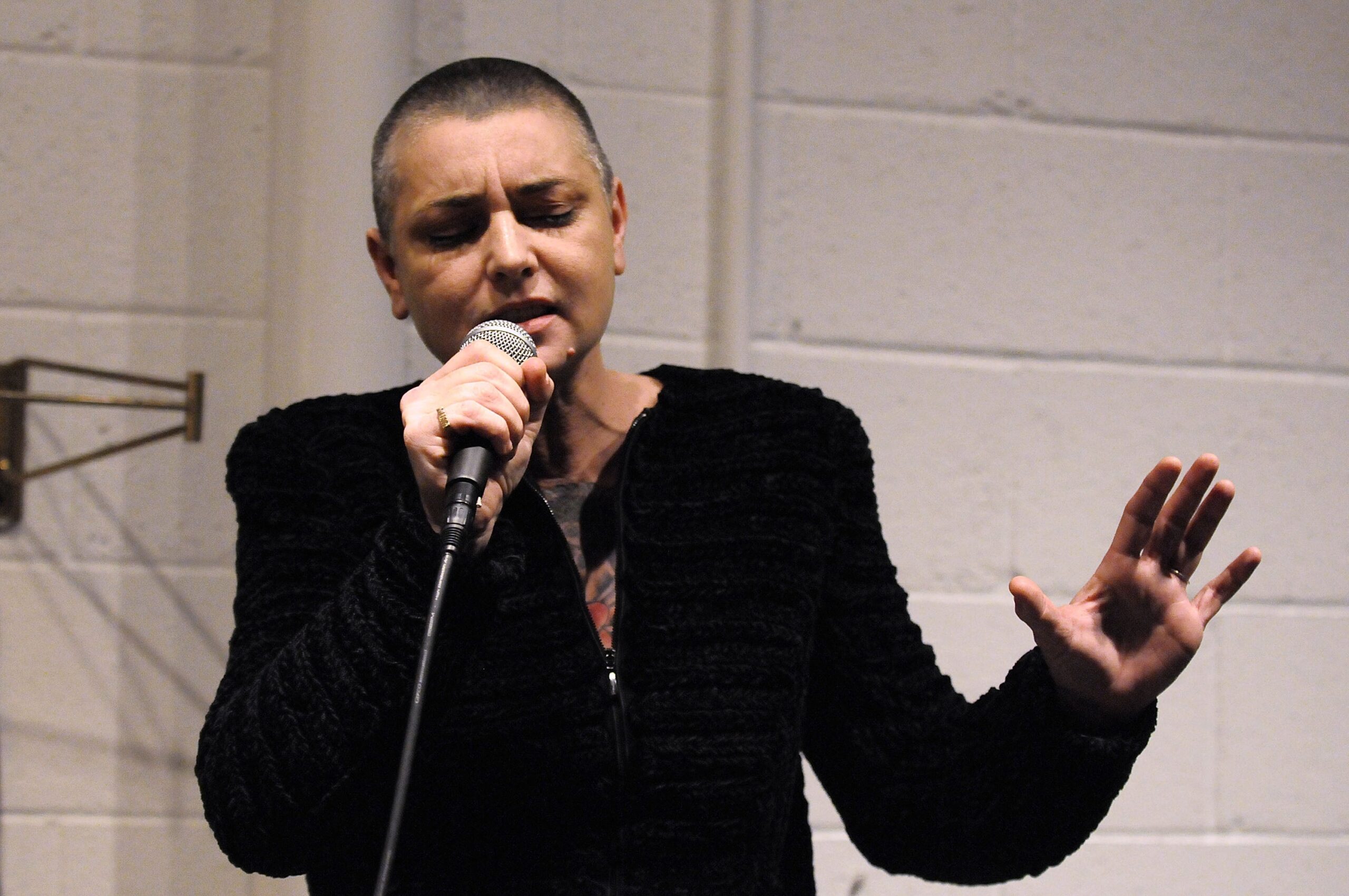 Sinead O'Connor with a shaved head is passionately singing into a microphone, wearing a textured jacket in front of a plain wall