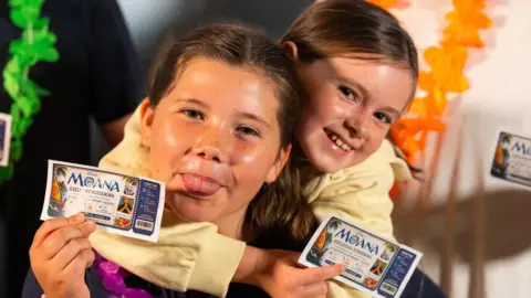 James Gillham/Disney A close-up image of two girls holding 'Moana Exclusive Screening' cinema tickets.  One is poking her tongue out. They are both smiling and looking into the camera.