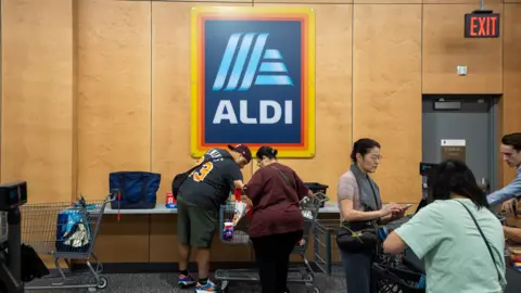 Aldi An Aldi logo sign hangs above a man in a blue jersey with yellow numbers, green shorts, and a backwards cap as he removes items from a cart and a woman with a burgundy shirt and black pants stands beside him. A woman in a pink shirt with a grey sweatshirt looks at her phone. Meanwhile, a woman with a green shirt removes items from a grocery cart.