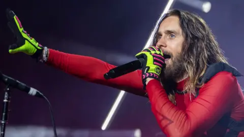 Getty Images Jared Leto stands under concert lighting, singing into a handheld microphone. He has long hair and a beard and is dressed in a red, sheer and fitted top with neon green and pink gloves. One arm is stretched outward toward the audience while the other holds the microphone close to his mouth. Bright white stage lights form diagonal lines in the background.CLARKSTON, MICHIGAN - AUGUST 06: Actor/Singer Jared Leto of Thirty Seconds To Mars performs during their Seasons World Tour 2024 at Pine Knob Music Theatre on August 06, 2024 in Clarkston, Michigan. (Photo by Scott Legato/Getty Images)
