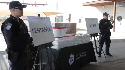 US Customs and Border Protection via Reuters A table laden with packets of white powder and two signs saying fentanyl and meth. Two men in uniform and baseball caps stand guard.