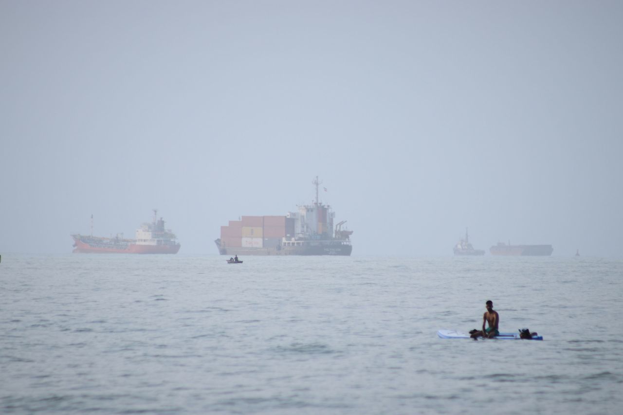 A man sits on a surfboard as a container ship and other commercial vessels appear anchored in the Strait of Hormuz off Bandar Abbas, Iran, Monday, July 27.