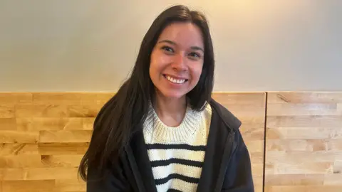 Montana State Representative and Bozeman local Katie Fire Thunder, 25, sits on a bench at a Bozeman coffee shop and smiles, her long dark hair flowing over a white top with black stripes and a black zip-up hoodie