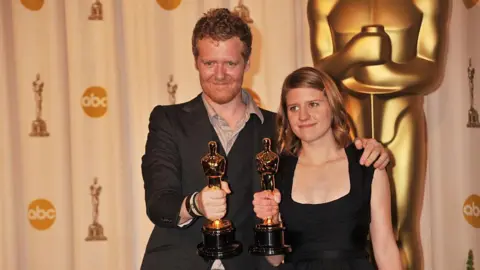 Getty Images Glen Hansard and Markéta Irglova hold their Oscars. Hansard has short red hair and stubble and is wearing a suit jacket and open-necked shirt. Irglova is wearing a black dress and has shoulder-length reddish/brown hair