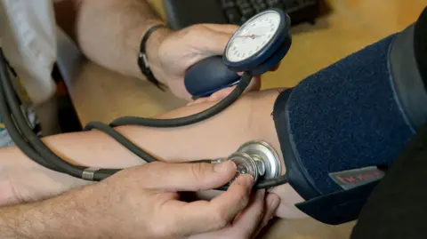 PA Media Close-up of a healthcare worker measuring a person's blood pressure using a cuff, gauge and stethoscope during a health check.