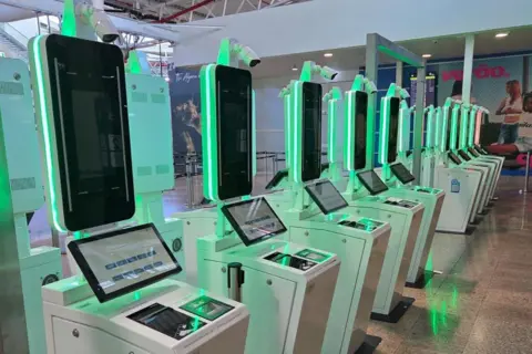 PSP / Pedro Oliveira A row of automated machines with screens and equipment to record facial scans, fingerprints and scan passports. They have green lighting.