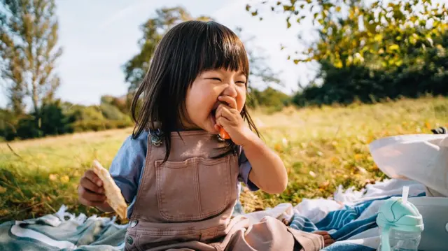 A little girl wey sidon outside dey eat carrot while holding a piece of bread for her oda hand.