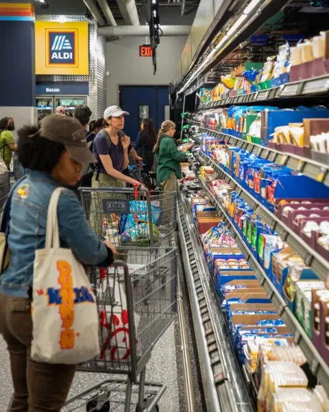 ALDI A woman in a jean shirt with brown jeans and a brown hat looks at cheese options while holding onto her shopping cart. Another woman in the background wearing green pants, a blue shirt and a beige hat looks at other dairy options, including a third woman in a green shirt, khaki pants looks at the labeling of an item.