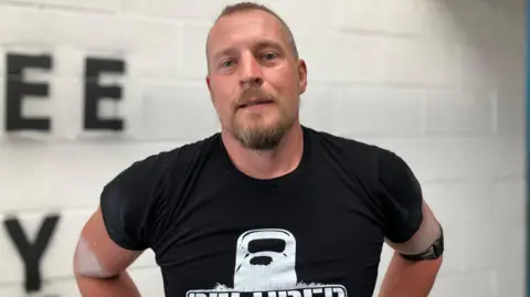 BBC A man with short hair and a short beard and moustache is looking at directly at the camera. He wears a black t-shirt with an outline of a kettlebell on it