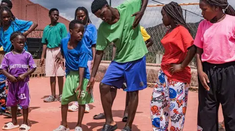 AFP via Getty Images Members of the Ugandan dance group Ghetto Kids rehearse at their home in Nakawuka, Uganda, on 25 May 2026. They are wearing an array of brightly coloured T-shirts and mimicking the moves of an adult man choreographer.
