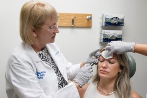 A physician injects Botox below the eyebrow of a woman patient. The hand of an assistant holds a piece of gauze on the patient’s forehead.