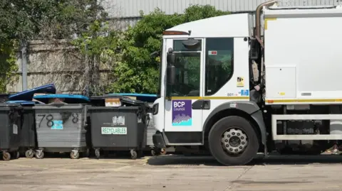 BBC A bin lorry next to some skips which are full.