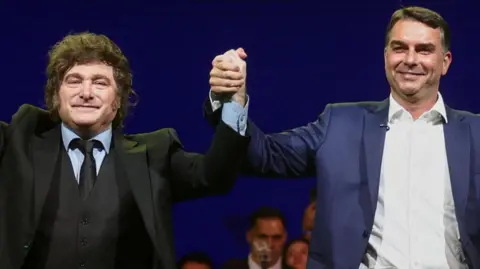 Reuters Argentine President Javier Milei holds hands aloft with Brazilian Senator Flavio Bolsonaro. Both men are wearing suits and smiling as they appear at a campaign event.
