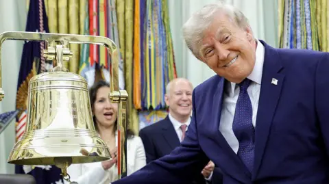 Getty Images President Donald Trump ringing the opening bell in the Oval Office on Monday. The US president is smiling at the cameras as he rings the big gold bell. He is wearing a dark blue suite, with a dark blue spotted tie and a white shirt.