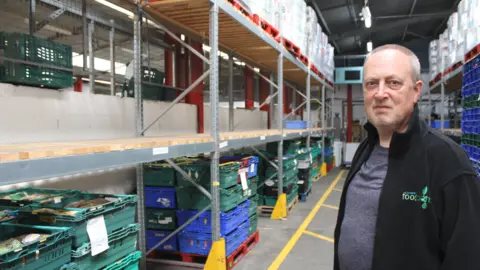 Coventry Foodbank A man with grey hair and a black jacket stands in a warehouse. Rows of empty shelves and stacks of crates can be seen behind him.