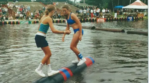 Samantha LaSalle Two women with their dirty blonde hair pulled back, one in a two-piece swimsuit and the other in shorts and a tank top, balance in socks and sneakers on a rolling log in water as stands of spectators look on