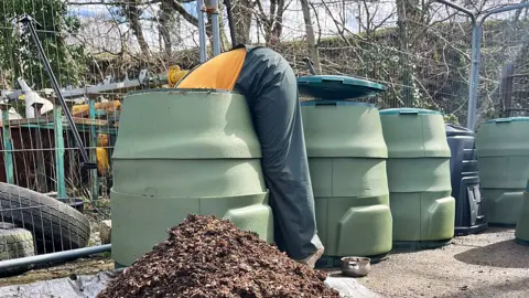 HeleddWyn Somebody wearing an orange t-shirt and green work overalls is bent over leaning a long way into a green composting or storage bin. There is a pile of compost in front of the bin. Three others stand to the right of it. Behind is movable metal fencing with tyres and other industrial type equipment behind it, and a tree and scrub covered embankment.
