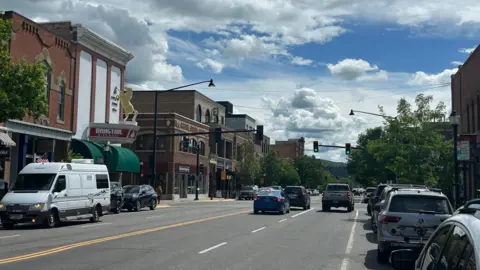 Cars and trucks drive under a blue sky along the main street of Bozeman, lined on either side by parked vehicles and two- or three-storey brick and Western-style buildings housing shops and restaurants