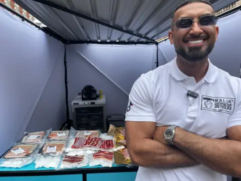 Manny Malik to the right of the shot wearing a white polo shirt with black Malik Butchers branding. To the left of the shot behind him is a selection of packed meats